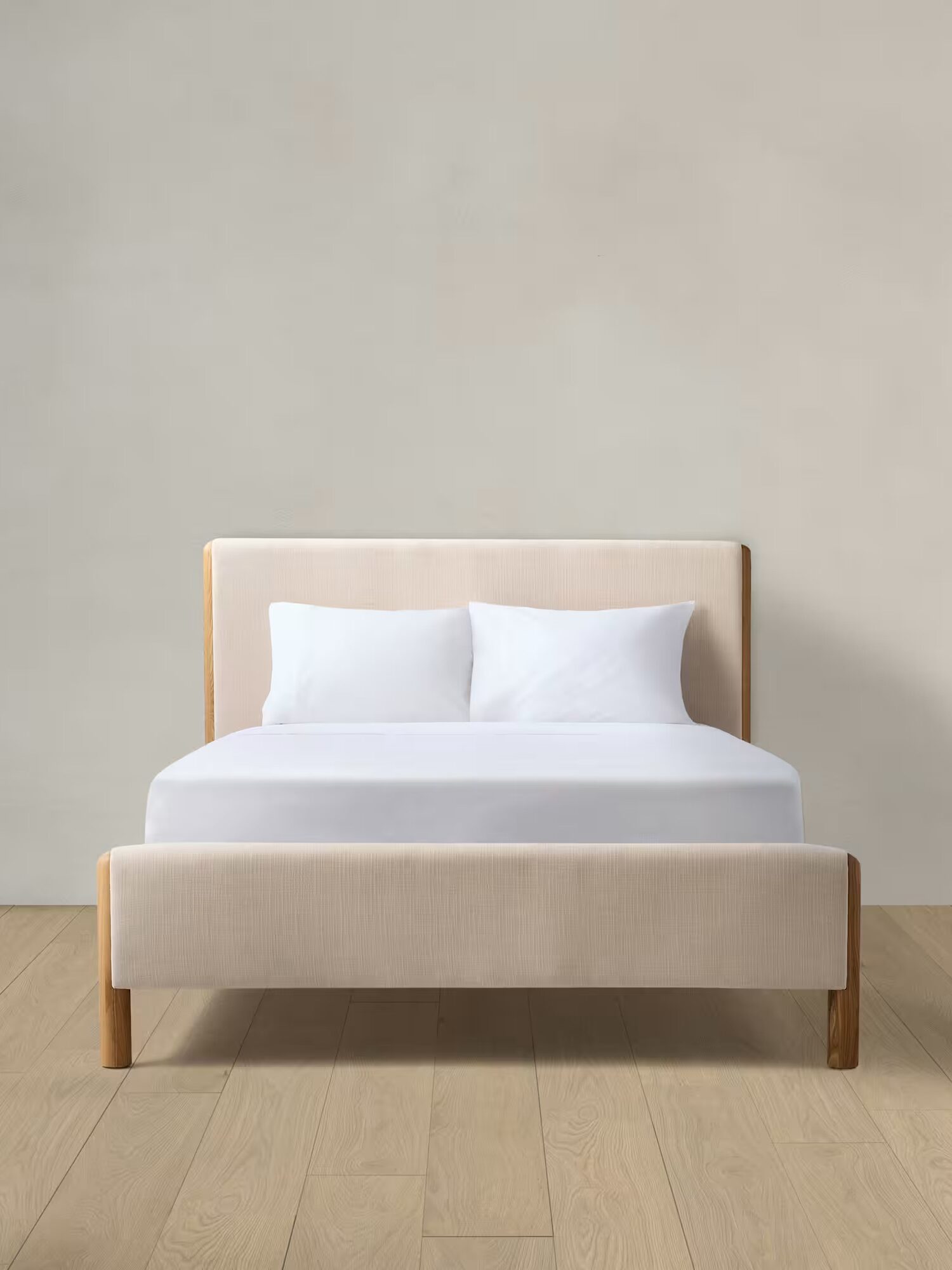 A beige upholstered bed with white bedding and two pillows is centered against a plain light-colored wall on a wooden floor.
