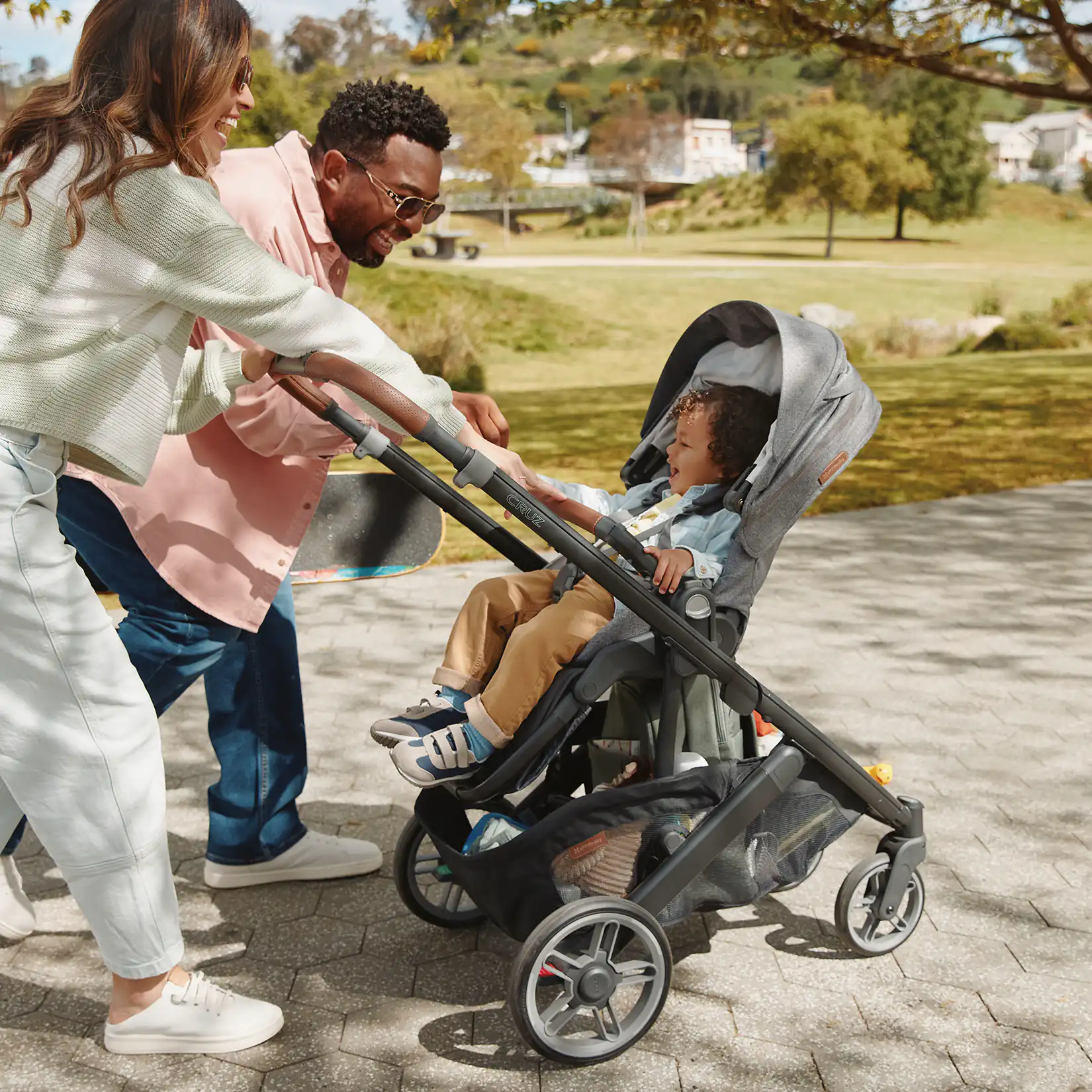 Two adults smile and interact with a young child sitting in a stroller outdoors on a sunny day in a park-like setting.