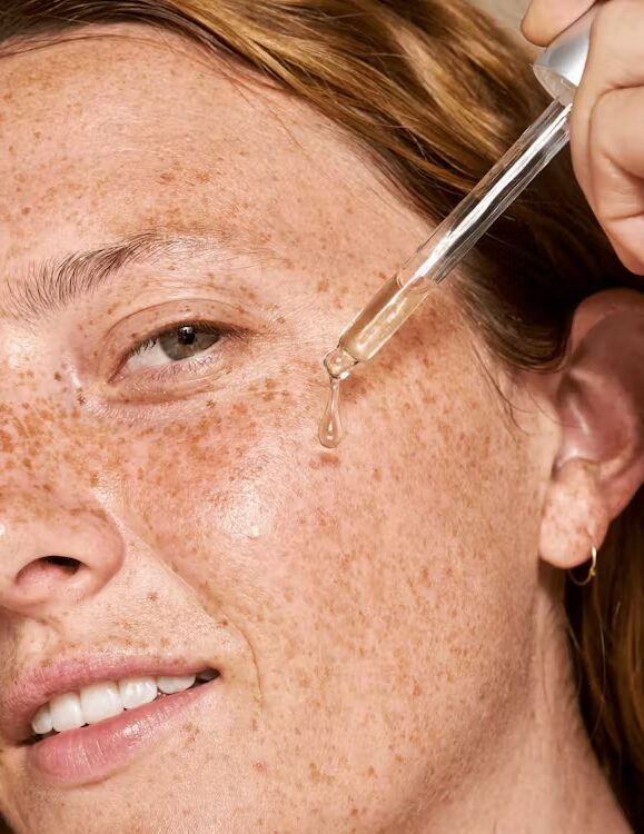 A person with freckles applies facial serum to their cheek using a dropper.
