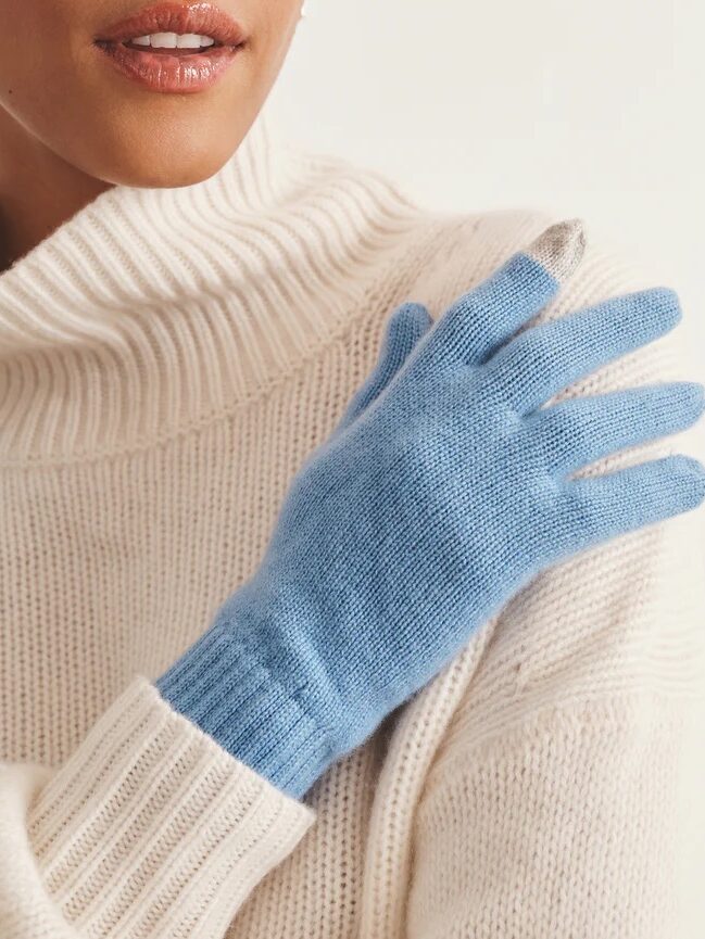 A person wearing a white ribbed turtleneck sweater and light blue knit glove rests their gloved hand on their chest.