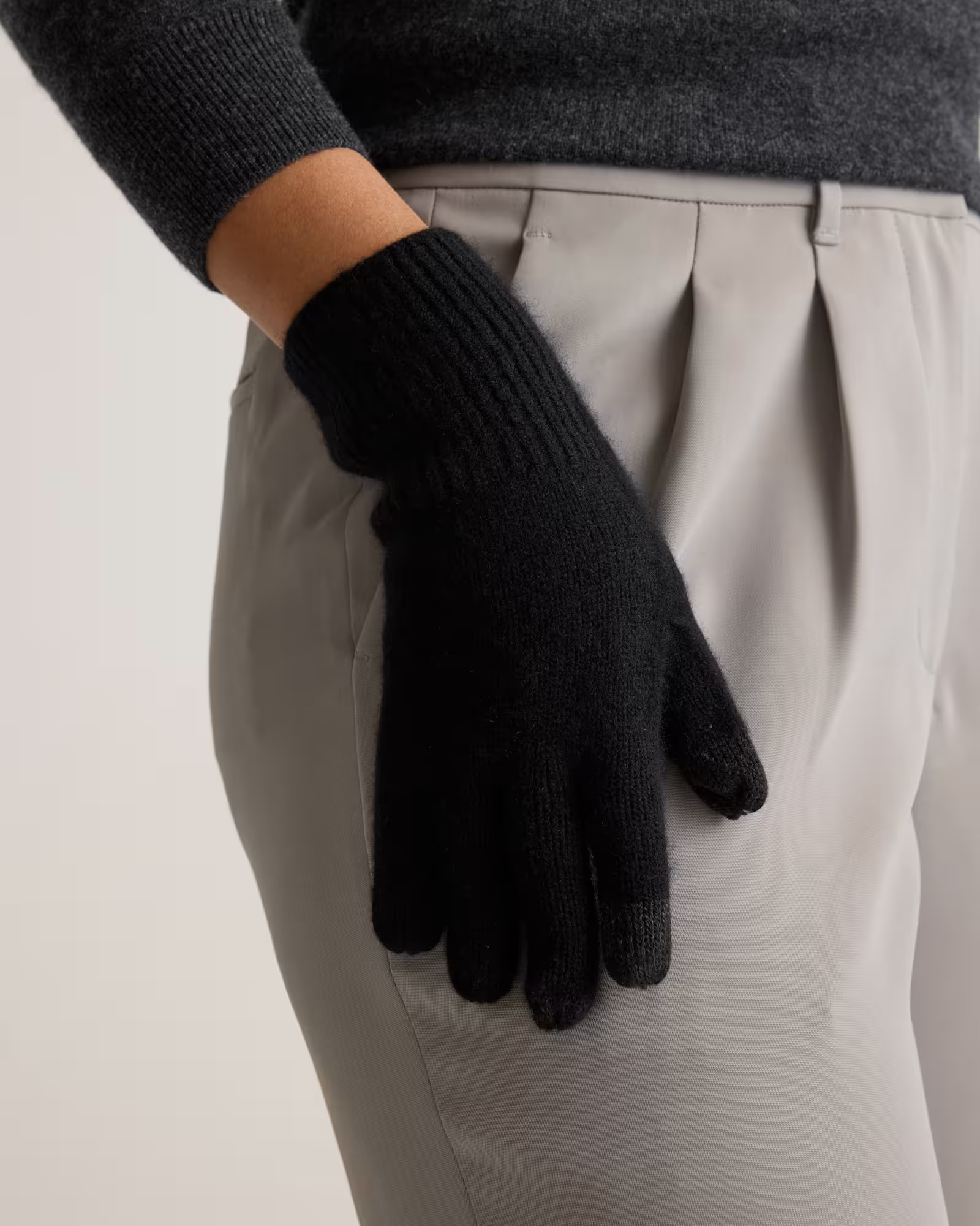 Person wearing a dark gray sweater and black gloves, with one hand resting on the side of light gray pleated pants.