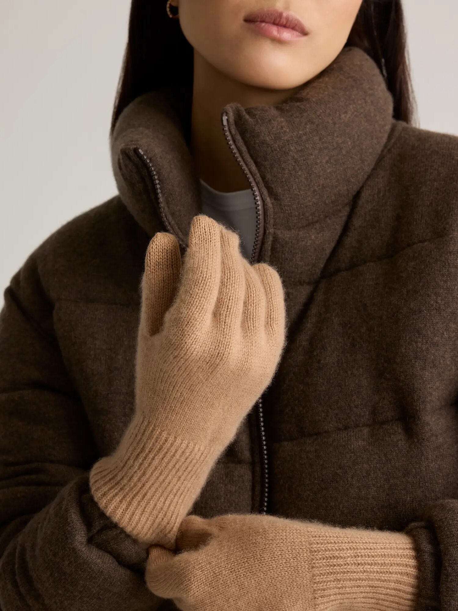 A woman wearing a brown zip-up coat and beige wool gloves, with one hand adjusting the zipper.