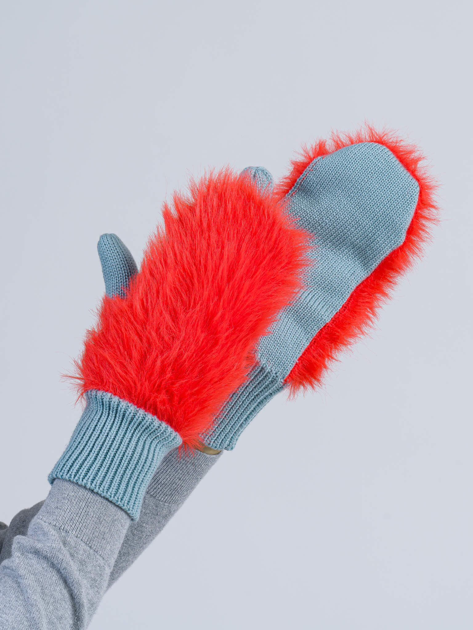 A person wearing gray sleeves holds up blue mittens with bright red faux fur on the backs against a plain light background.