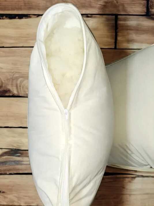 A white pillow with a partially open zipper shows wool filling inside. Text above highlights features: 100% cotton outer layer, 100% merino wool filling, and handmade with care.