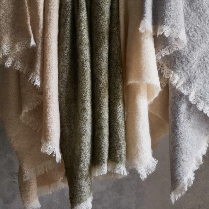Several fringed, textured fabrics in neutral tones of beige, gray, and green hang vertically against a plain background.