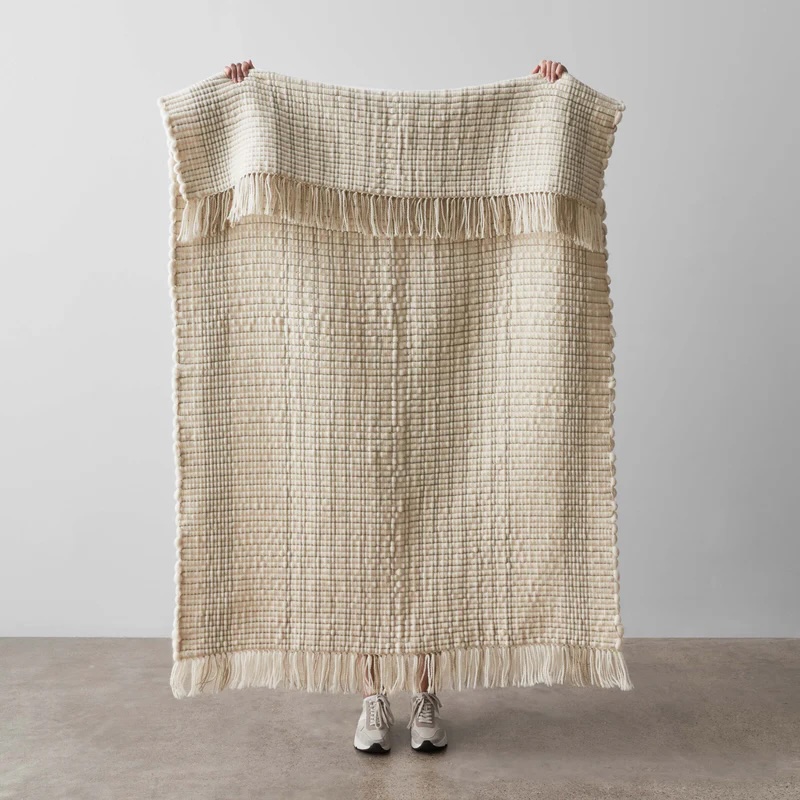 A person stands behind a cream-colored, textured woven blanket with fringe, holding it up fully extended; only their hands and shoes are visible.