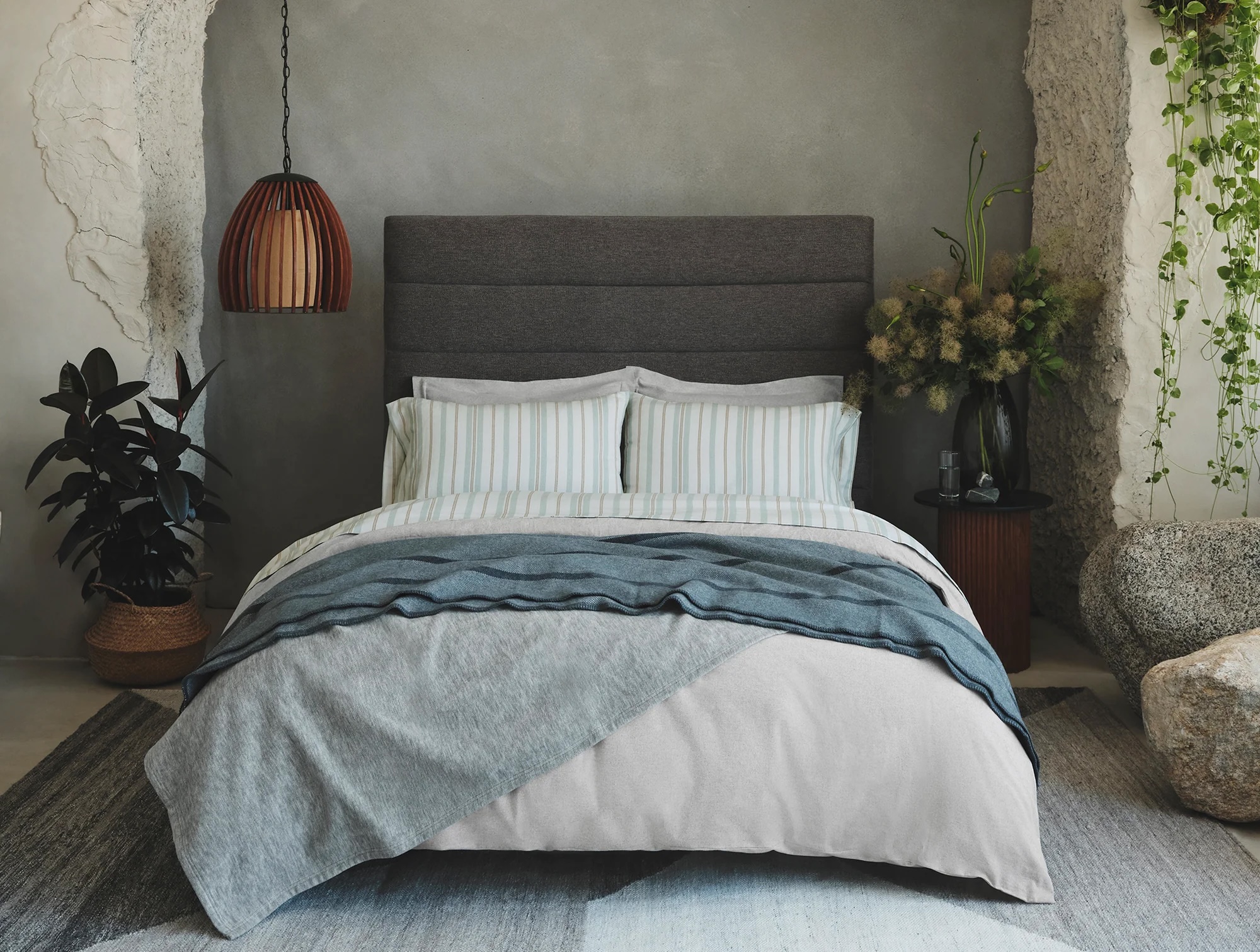 A modern bedroom with a dark upholstered headboard, neatly made bed with layered bedding, plants, a pendant lamp, and textured gray walls.