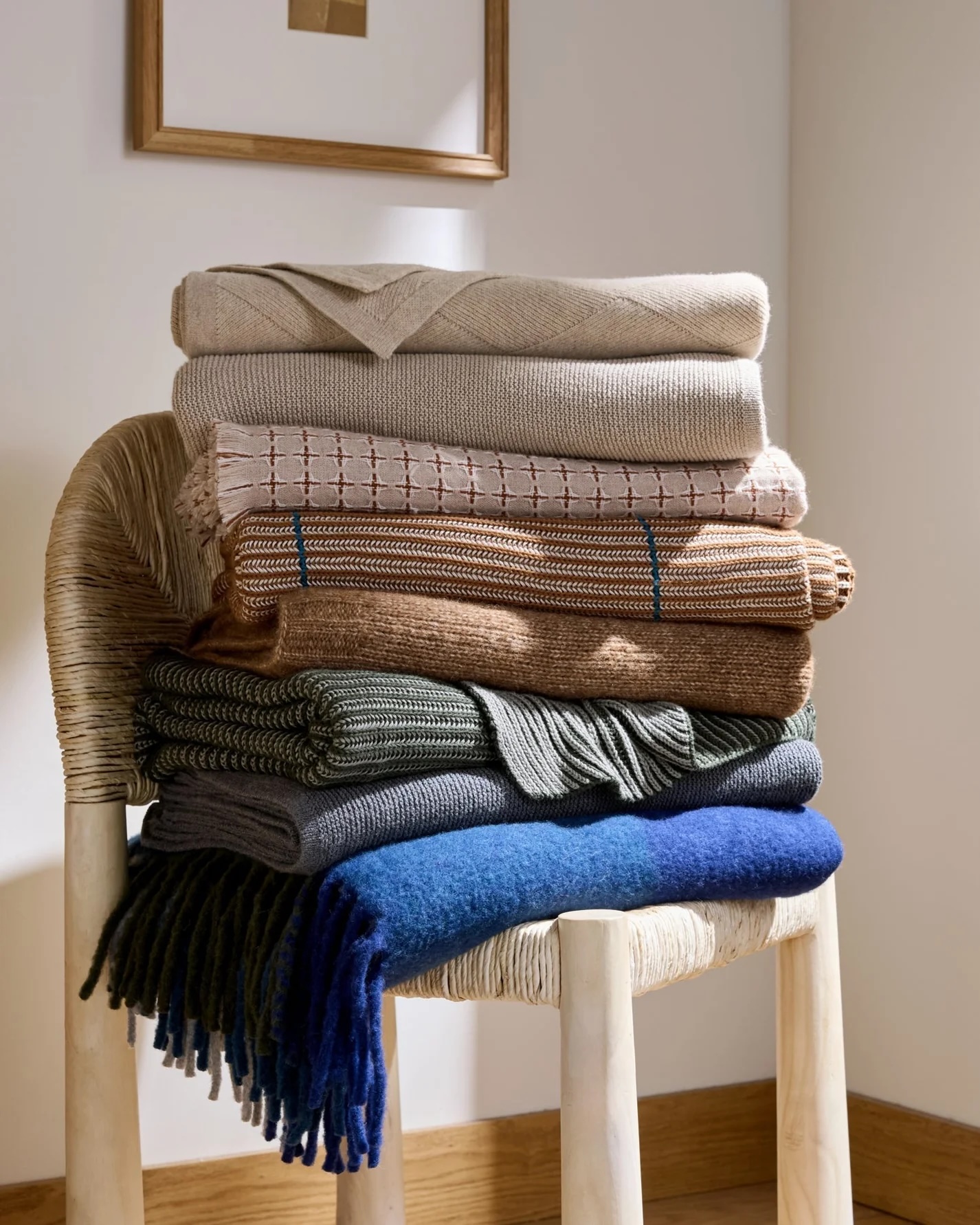 Six folded blankets in beige, brown, green, gray, and blue are stacked on a woven chair in a softly lit room with light wood flooring.