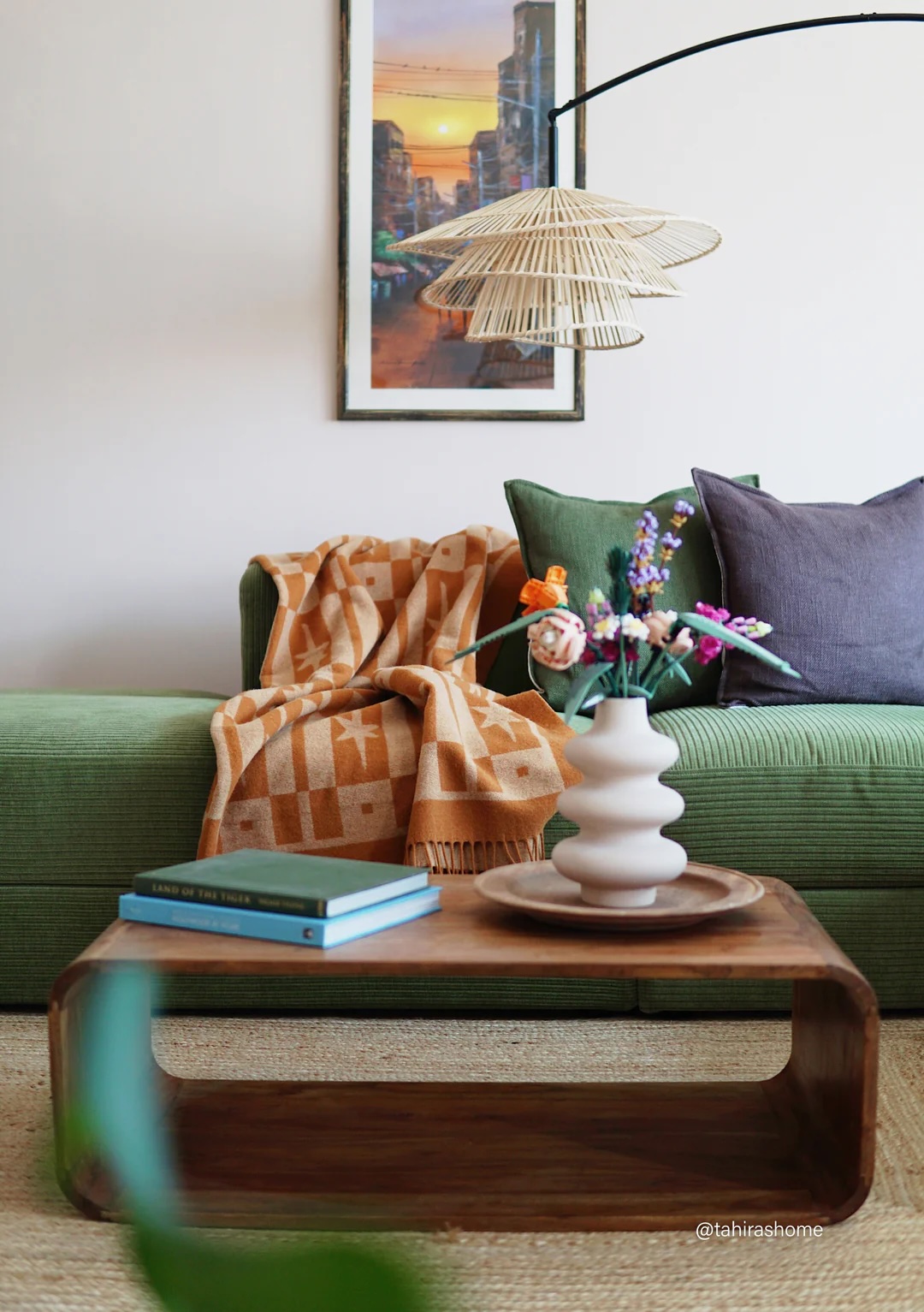 A green sofa with colorful cushions and an orange patterned blanket sits behind a wooden coffee table with a vase of flowers, books, and a tray. A framed artwork and pendant light are above.