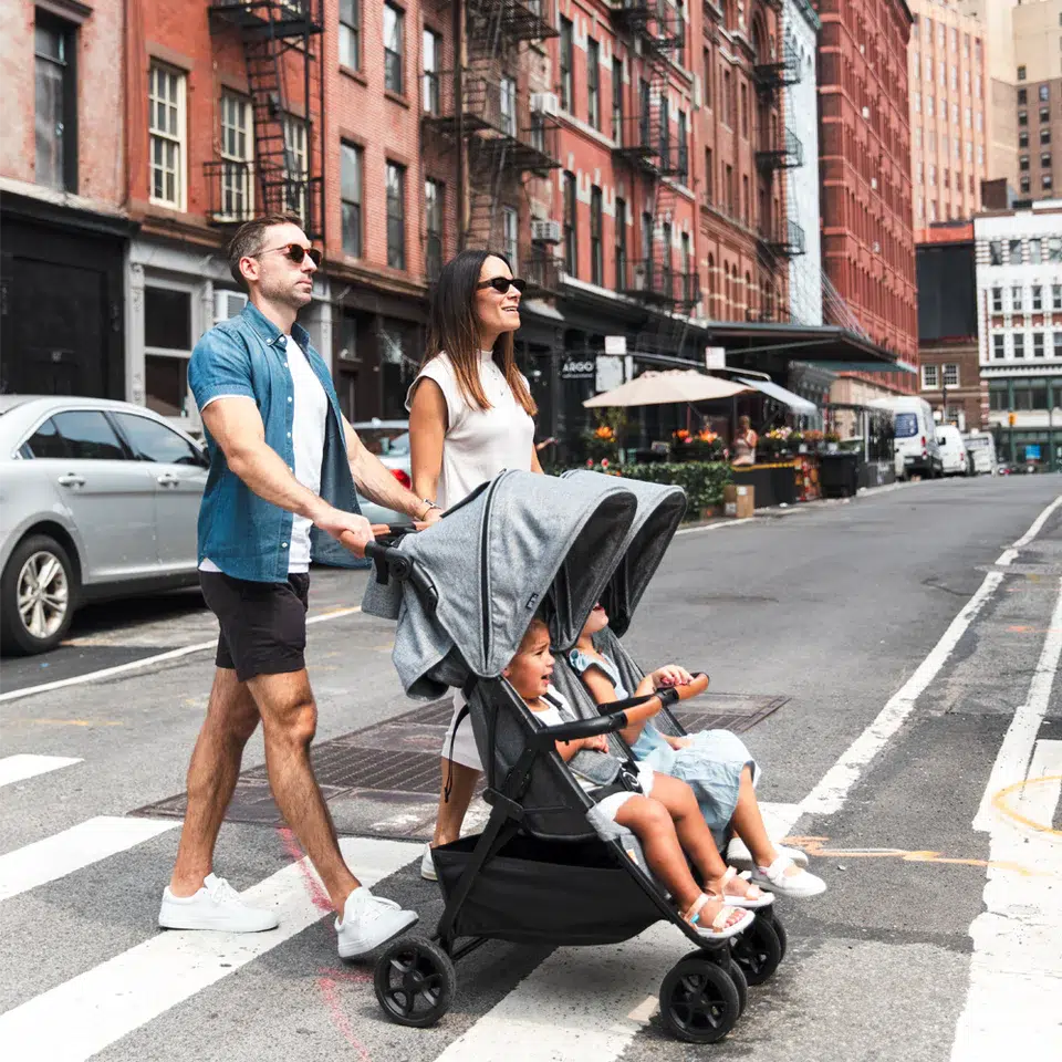A man and woman push a double stroller with two children across a city street at a crosswalk on a sunny day.