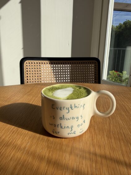 A cup of green matcha latte with heart-shaped foam sits on a wooden table. The mug reads, "Everything is always working out for me :)" A window and chair are in the background.