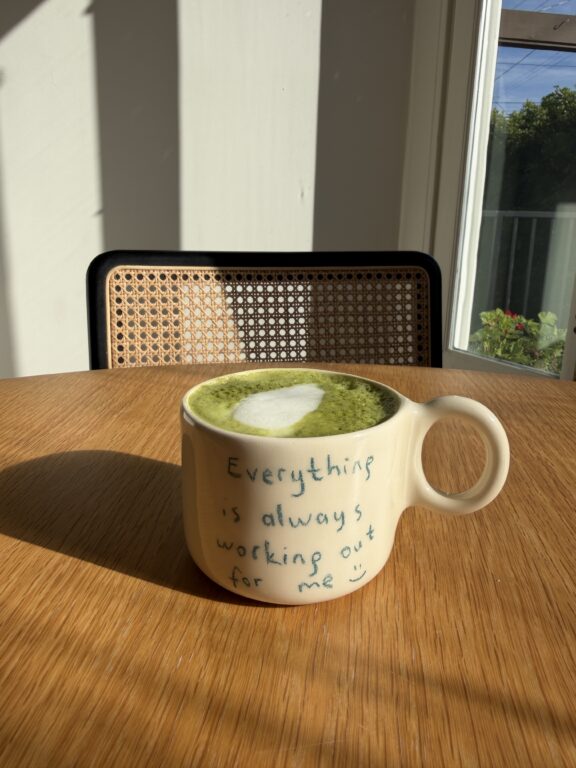 A cup of green matcha latte with heart-shaped foam sits on a wooden table. The mug reads, "Everything is always working out for me :)" A window and chair are in the background.