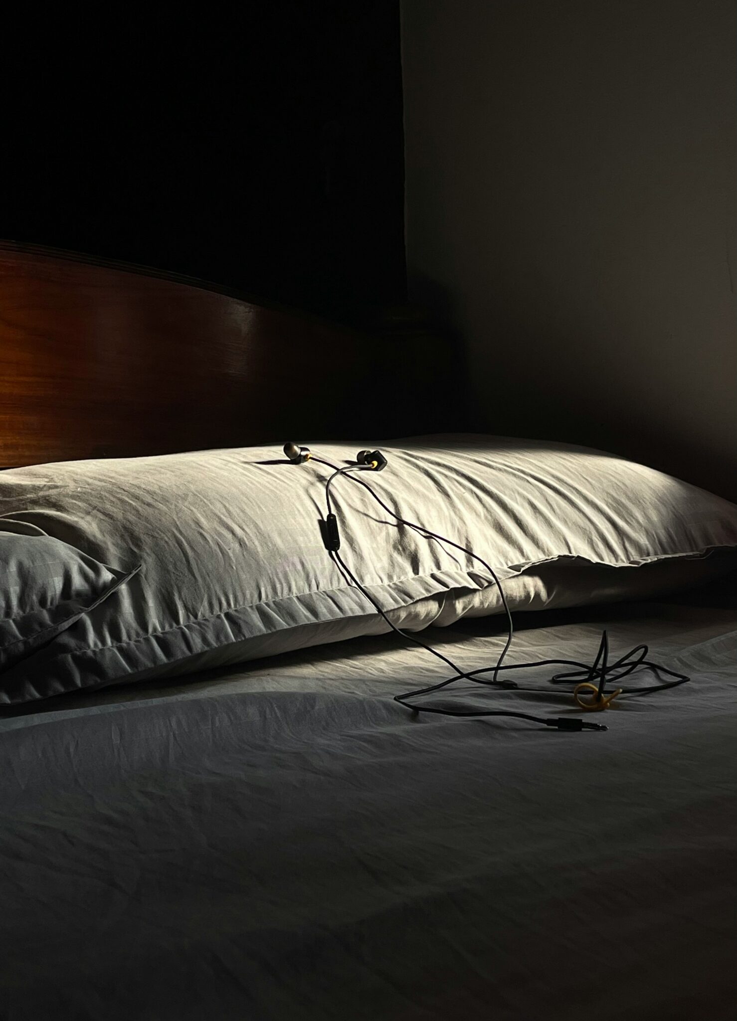 A pair of wired earphones rests on a white pillow on an unmade bed, with sunlight casting shadows across the scene.