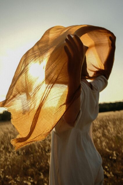 Person in a white outfit stands in a field, holding a translucent brown scarf overhead as sunlight shines through the fabric.