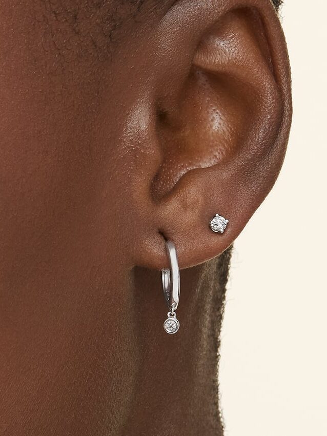 Close-up of an ear wearing two earrings: a small round stud in the upper lobe and a silver hoop with a small dangling gem in the lower lobe.