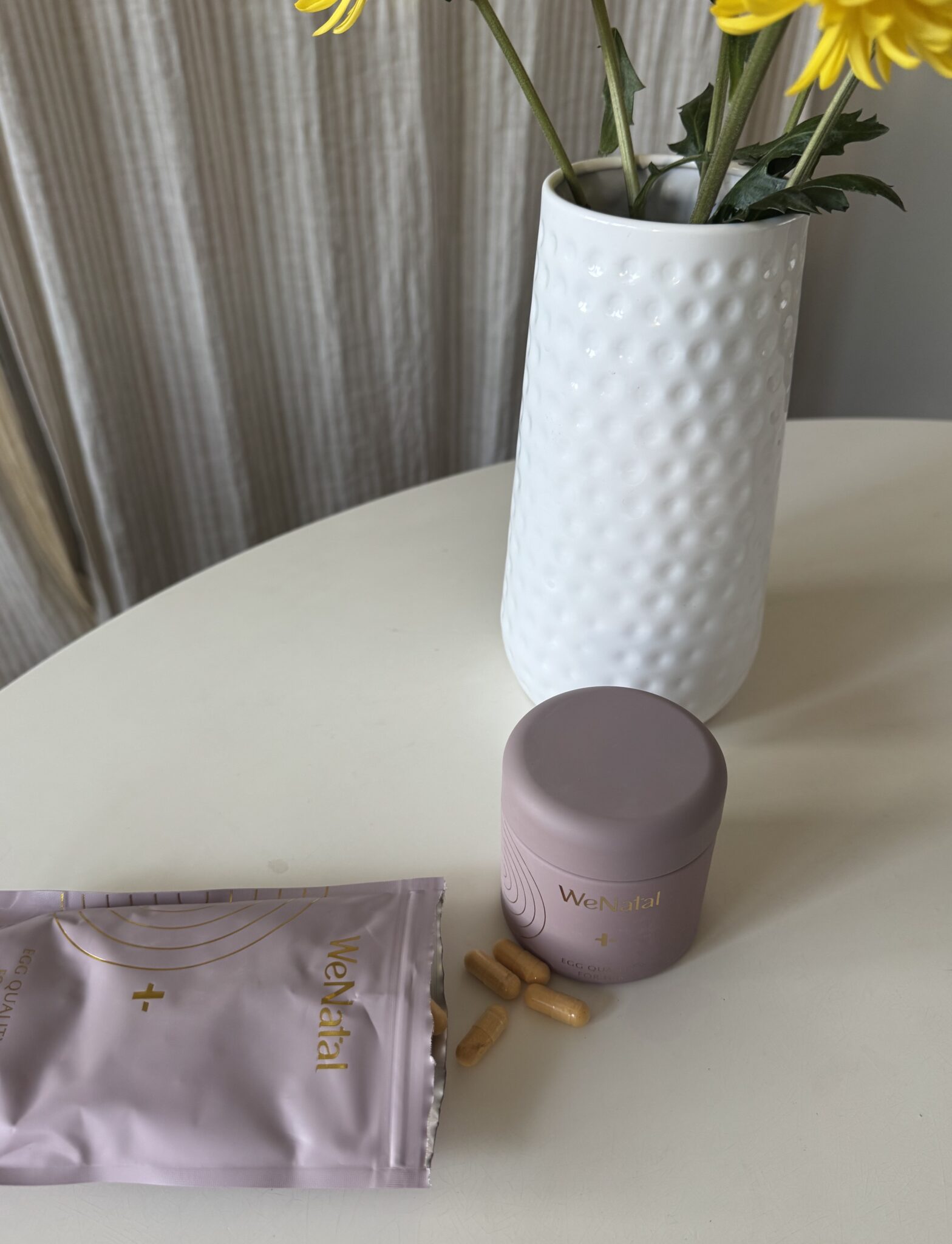 A mauve container and a matching pouch labeled "Welltai" are on a round white table with several yellow capsules beside them and a white vase with yellow flowers in the background.