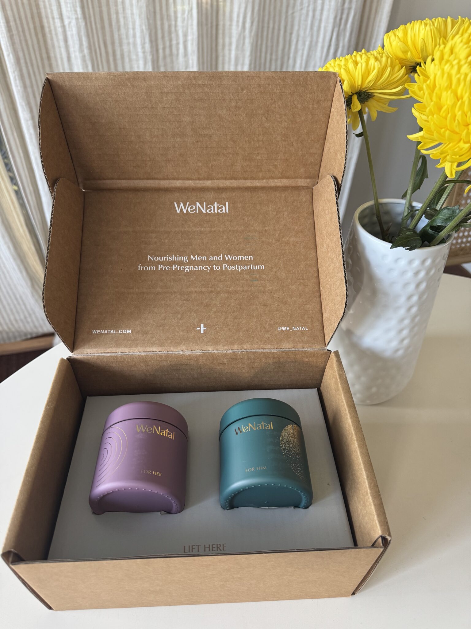 Two WeNatal supplement containers in a cardboard box on a white table, with a vase of yellow flowers in the background.