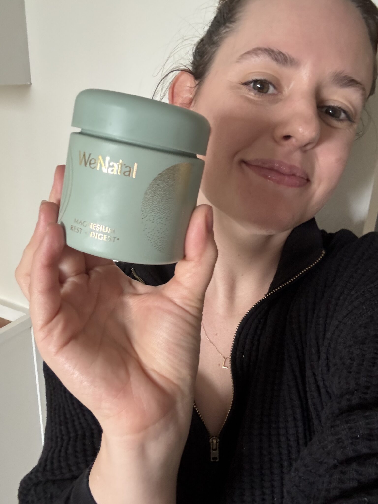 A woman in a black top holds up a green container labeled "WeNatal" with gold lettering and smiles at the camera.