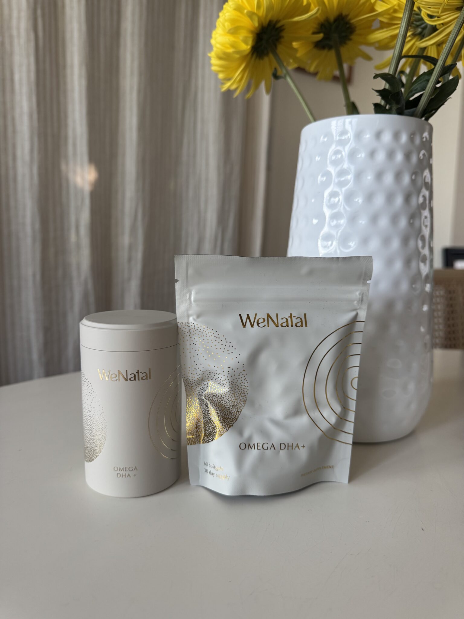 Two WeNatal Omega DHA+ supplement containers, one cylindrical and one resealable pouch, are displayed on a white table next to a white textured vase with yellow flowers.