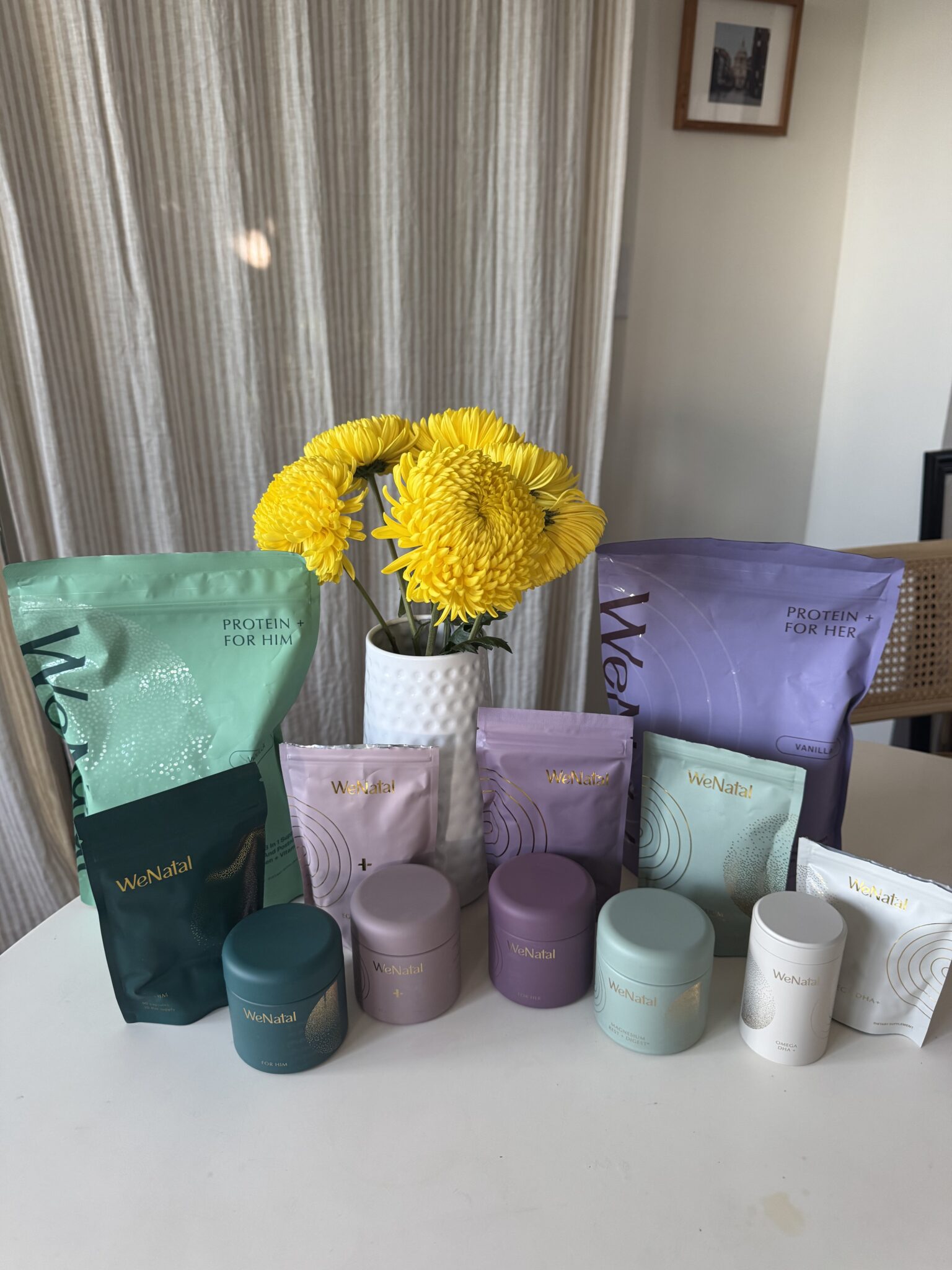 A display of various WeNatal supplement containers and pouches arranged on a white table with a vase of yellow flowers in the center. A curtain and framed photo are in the background.