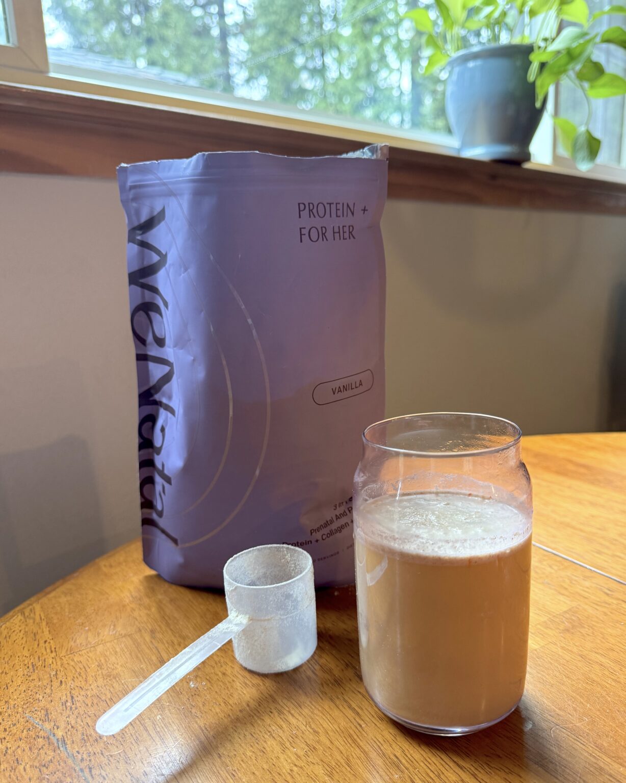 A bag labeled "Protein + For Her Vanilla," a scoop, and a glass of mixed protein shake sit on a wooden table near a window with a potted plant in the background.