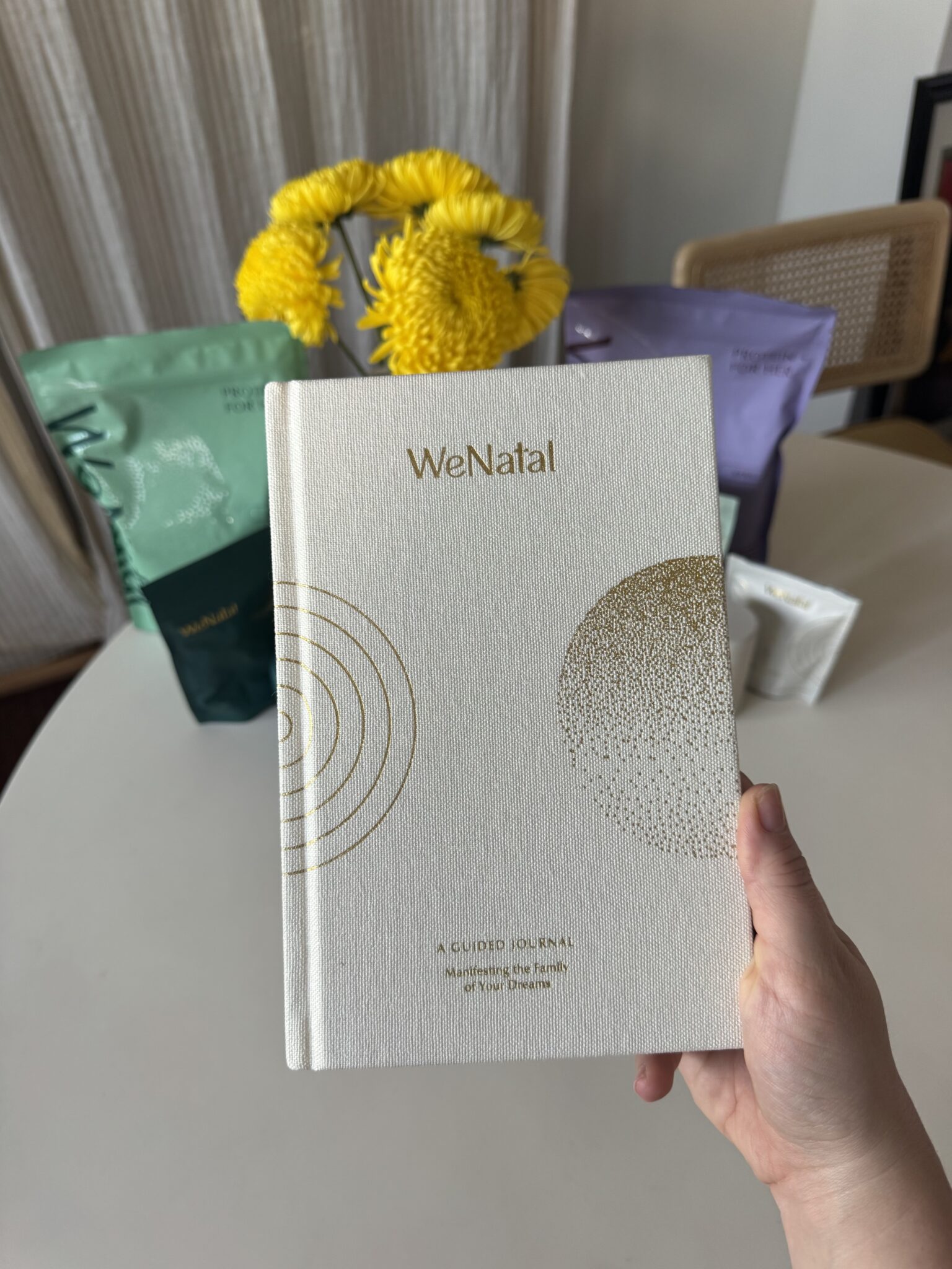 A hand holds a white WeNatal journal with gold designs. In the background, yellow flowers and green and purple packets are on a white table.