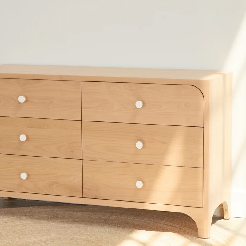 A light wood dresser with six drawers and white round knobs sits against a white wall in natural sunlight.