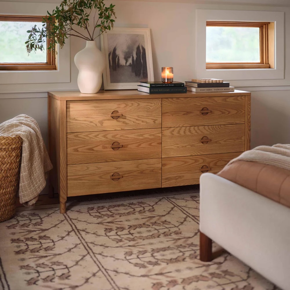 A wooden dresser with six drawers is topped with books, a candle, a white vase with branches, and a framed picture. The dresser sits next to a bed in a softly lit room.