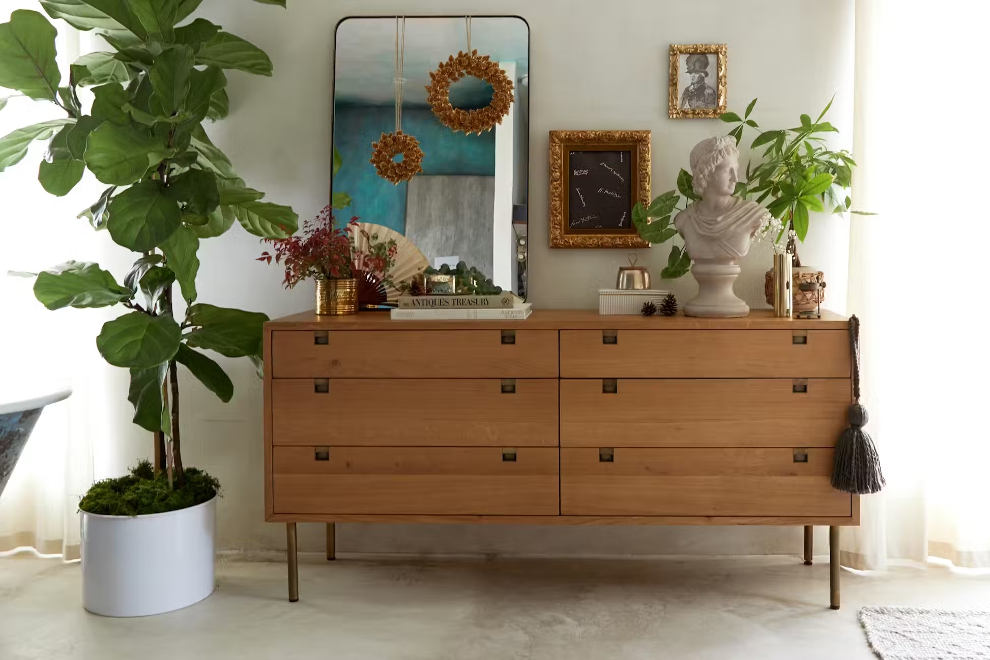 Wooden dresser with brass handles topped with plants, books, and decorative items, positioned against a wall with framed art and a tall mirror; potted plant beside dresser.