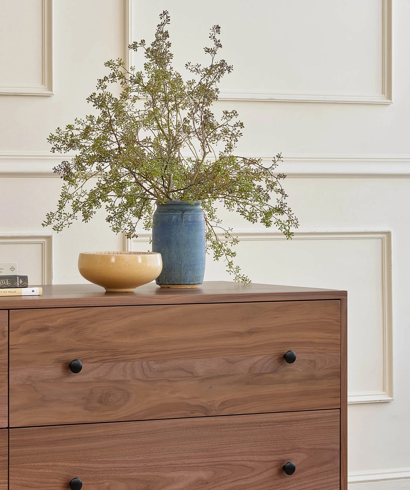 A wooden dresser with black knobs holds a blue vase with leafy branches and a yellow bowl, set against a white paneled wall.