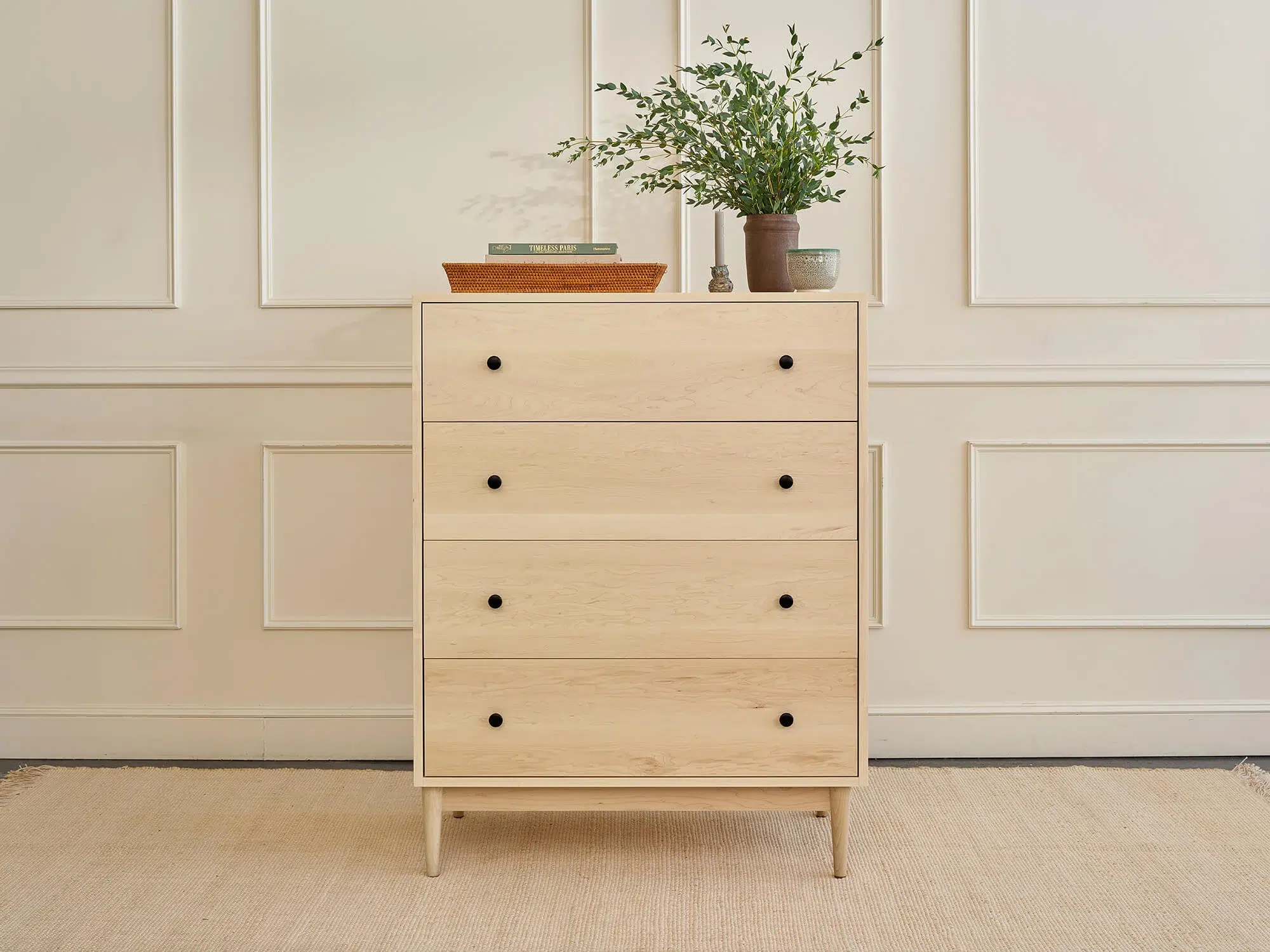 Light wood dresser with four drawers and black knobs stands against a beige paneled wall. A plant, a stack of books, and a tray decorate the top. A neutral rug covers the floor.