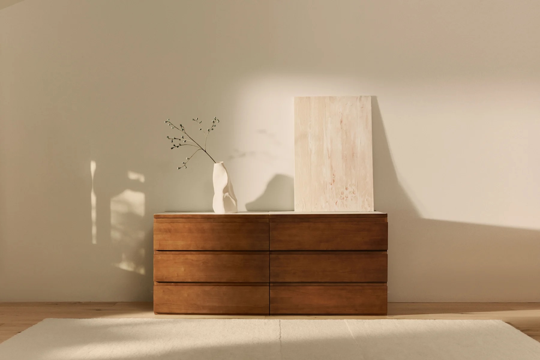 A wooden dresser with six drawers stands against a beige wall, topped with a white vase holding a branch and a rectangular textured artwork.