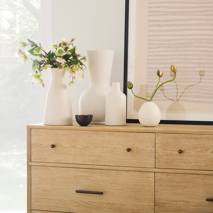 A wooden dresser with four drawers holds white ceramic vases, one with flowers, a small black bowl, and a large framed abstract artwork leaning against the wall.