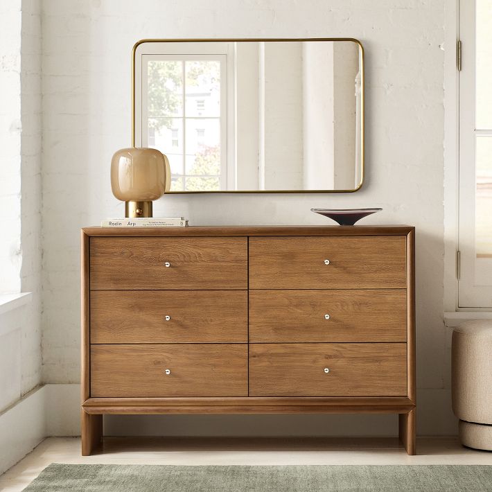 A wooden dresser with six drawers stands against a white wall, topped with a lamp, books, and a dish, with a rectangular mirror hanging above it.
